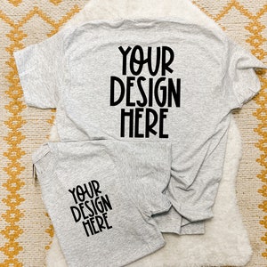 May include: Two gray t-shirts with the text "YOUR DESIGN HERE" printed on them. The shirts are folded and laid out on a white fuzzy surface.