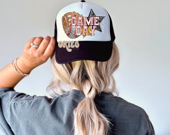 May include: Black and white trucker hat with a white mesh back. The front of the hat features a glittery design of a baseball glove, baseball, and a star with the text "GAME DAY" in red and white.