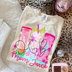 Pu&ograve; includere: Felpa Comfort Colors color crema con un design grafico con due lattine di bevande rosa e una tazza. Il testo sulla maglietta recita "Mom Juice" in corsivo rosa. Le lattine sono etichettate "Strawberry Sunrise".