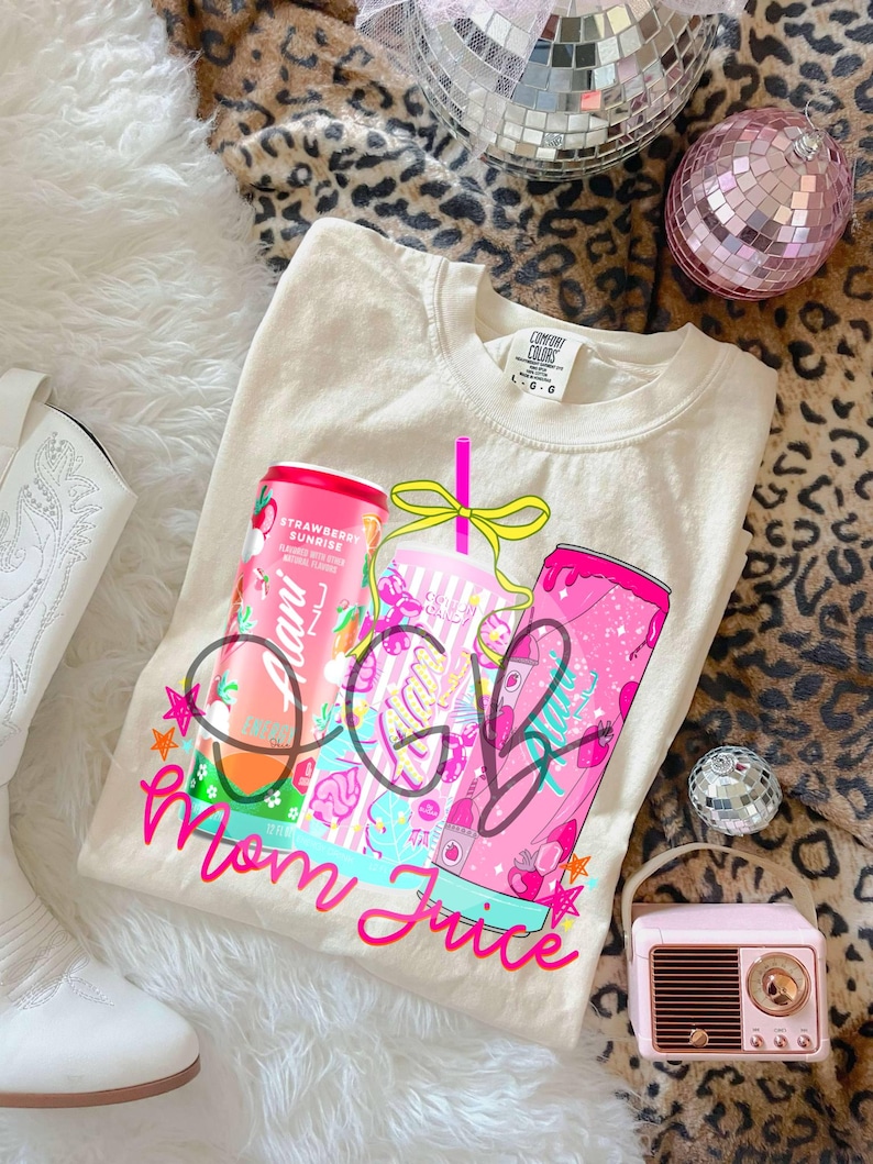 Pu&ograve; includere: Felpa Comfort Colors color crema con un design grafico con due lattine di bevande rosa e una tazza. Il testo sulla maglietta recita "Mom Juice" in corsivo rosa. Le lattine sono etichettate "Strawberry Sunrise".