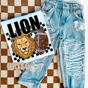 Könnte beinhalten: Weißes T-Shirt mit einem schwarz-weiß karierten Hintergrund. Das Shirt zeigt einen Comic-Löwen mit einem Fußball und dem Text "Lion Pride".