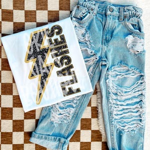 May include: A white t-shirt with a black and gold lightning bolt graphic and the text "FLASHES" in black glitter. The shirt is paired with light blue distressed denim jeans.