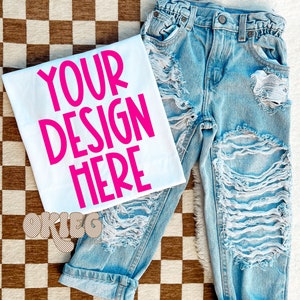 May include: White t-shirt with the text "YOUR DESIGN HERE" in pink lettering. The t-shirt is lying on a checkered brown and white surface with a pair of light blue distressed denim jeans.