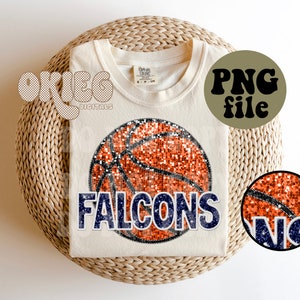 Puede incluir: Una camiseta blanca con una pelota de baloncesto naranja brillante y la palabra "FALCONS" en letras azules brillantes. La pelota de baloncesto tiene líneas negras y un contorno negro. La camiseta está sobre una cesta de mimbre.