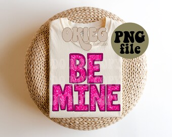 May include: A white t-shirt with the words "BE MINE" in pink glitter letters. The letters are in a retro font style.