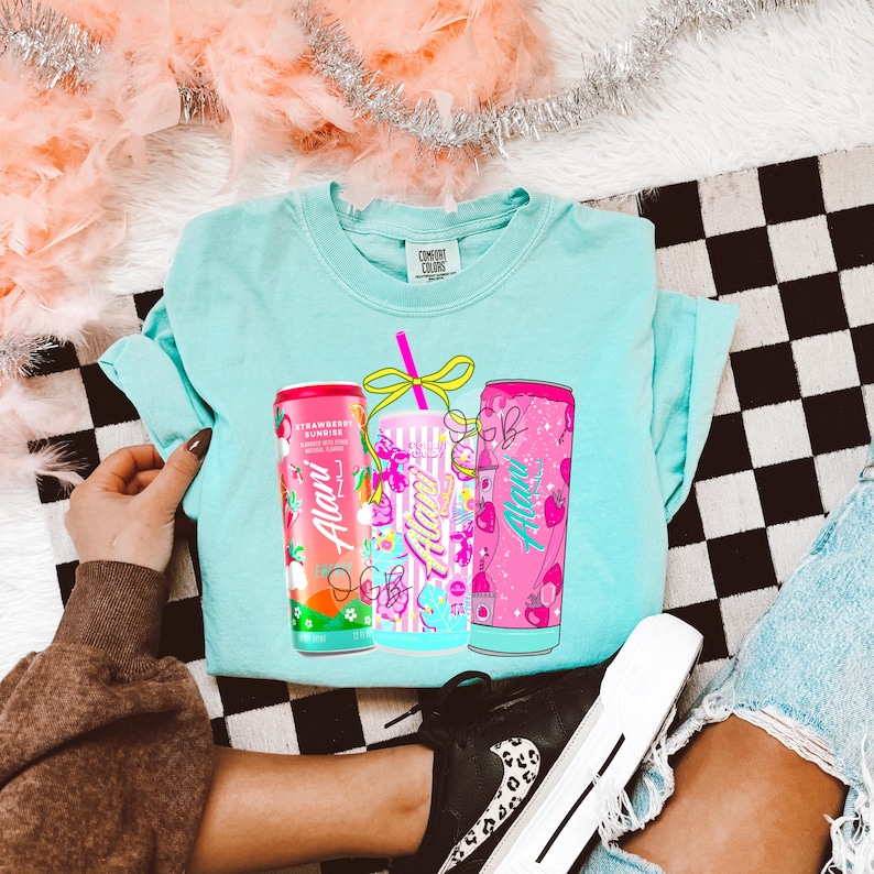 Op de afbeelding: Een lichtblauw T-shirt met een afbeelding van drie drankblikjes met de tekst "Alani". De blikjes zijn in roze, blauw en rood, met aardbeien- en fruitontwerpen. Het shirt ligt op een zwart-wit geblokt oppervlak.