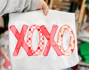 Puede incluir: Sudadera blanca con un gráfico "XOXO" rojo, rosa y blanco. Las letras están formadas por diferentes patrones, incluyendo cuadros, corazones y lazos.