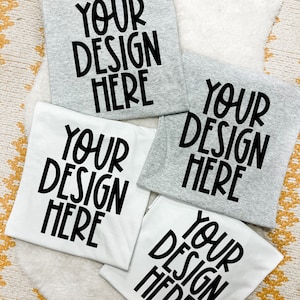 May include: Four folded t-shirts, two gray and two white, are arranged on a white fluffy surface. Each shirt has the text "YOUR DESIGN HERE" printed in bold black letters, offering a customizable design option.