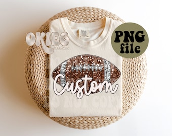Könnte beinhalten: Ein weißes T-Shirt mit einem braunen und silbernen Glitzer-Fußball-Design und dem Wort "Custom" in weißer Schrift. Das Shirt liegt auf einer geflochtenen Strohmatte.