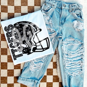 Könnte beinhalten: Ein weißes T-Shirt mit einer schwarz-weißen Grafik eines Footballhelms und dem Text "TIGERS" darauf. Das T-Shirt liegt auf einer braunen und weißen karierten Oberfläche mit einer hellblauen, ausgefransten Jeans.