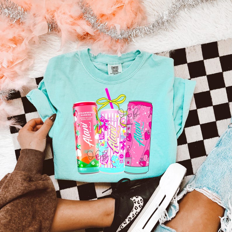 Op de afbeelding: Een lichtblauw T-shirt met een afbeelding van drie drankblikjes met de tekst "Alani". De blikjes zijn roze, blauw en rood, met aardbeien- en fruitontwerpen. Het shirt ligt op een zwart-wit geruit oppervlak.