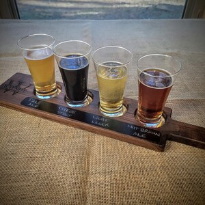 May include: A wooden beer flight holding four glasses of beer, each a different shade. The beers are labelled "Pale Ale", "Coffee Stout", "Light Lager", and "Nut Brown Ale". The flight is on a hessian surface, with a window in the background.