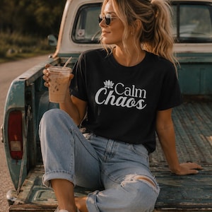 Puede incluir: Camiseta negra con las palabras "Calm Chaos" en escritura blanca y un gráfico de flor de loto. La camiseta se combina con vaqueros desgastados de color claro y zapatillas blancas. La persona sostiene un vaso de plástico transparente con una pajita.