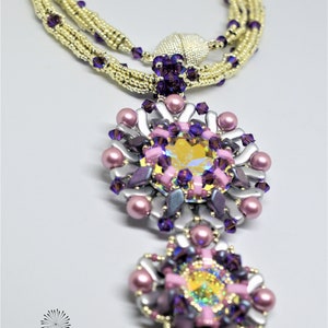 Op de afbeelding: Een meerstrengs ketting met een gouden en paarse kralenketting en een hanger gemaakt van zilver, roze en paarse kralen met een groot, iriserend kristal in het midden. De hanger heeft de vorm van een bloem. De ketting heet "Iris Necklace" en is van "Shrii Designs".