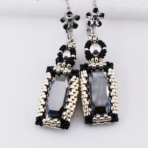 Puede incluir: Un par de pendientes de cuentas plateadas y negras con una piedra rectangular facetada de color gris oscuro. Los pendientes tienen un gancho en forma de flor y están etiquetados como "Diva Earrings (c) 2023 Silver Night/silver colorway".