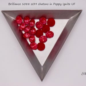 May include: A triangular metal tray contains several red chatons. The chatons are a mix of faceted and smooth, in a vibrant, poppy-red colour. The text "Brilliance 1088 ss39 chatons in Poppy Ignite UF" is at the top.