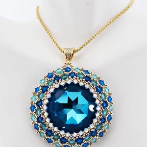 May include: A gold chain necklace with a blue and green beaded pendant featuring a large blue crystal. The pendant is round and has a multi-layered design. The text "Kumudha Pendant (c)2024 Blue/champagne" is visible above the pendant.