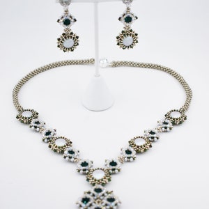 May include: Andromeda necklace and earrings set in emerald and light pewter. The necklace and earrings feature a beaded design with white, green, and silver accents. The earrings are displayed on a white stand. The necklace has a pendant with a teardrop-shaped emerald.