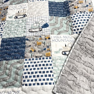 May include: A blue and gray patchwork quilt with a white and gray plush backing. The quilt features a repeating pattern of construction vehicles and the text "I will move mountains".