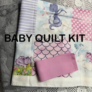 May include: A baby quilt kit with a gray plush blanket, a patchwork quilt top, and a pink ribbon. The quilt top features a dragon and cloud design, with pink and purple accents. The text "BABY QUILT KIT" is displayed.