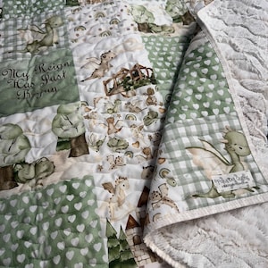 May include: A green and white baby quilt with a patchwork design featuring dragons, castles, and rainbows. The quilt has a soft, white minky backing and the text "My Reign Has Just Begun".