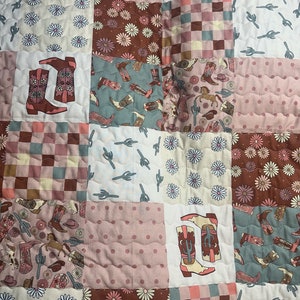 May include: A patchwork quilt with a variety of patterns, including floral designs, cactus prints, and cowboy boots. The quilt is made in shades of pink, brown, and blue.