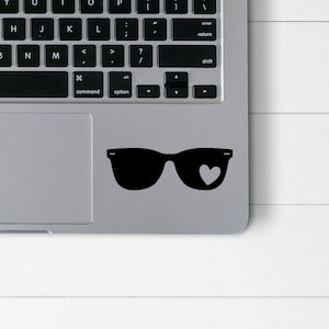 Puede incluir: Un ordenador portátil plateado con una pegatina de gafas de sol negras en el trackpad. Las gafas de sol tienen una forma de corazón en la lente.