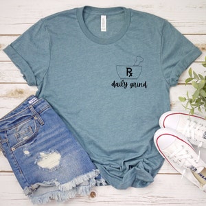 May include: A teal blue t-shirt with a black graphic of a mortar and pestle with an Rx symbol and the text "daily grind". The shirt is shown on a wooden surface with a pair of white sneakers and a pair of blue denim shorts.
