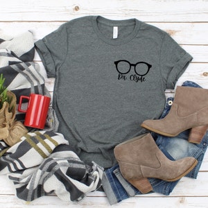 May include: A dark gray t-shirt with a black graphic of a pair of glasses and the text "Dr. Clyde".