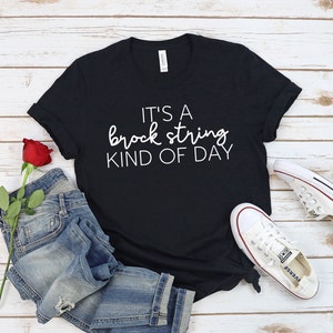Puede incluir: Camiseta negra con texto blanco que dice "It's a brock string kind of day". La camiseta está sobre una superficie de madera con un par de pantalones vaqueros azules y una rosa roja.