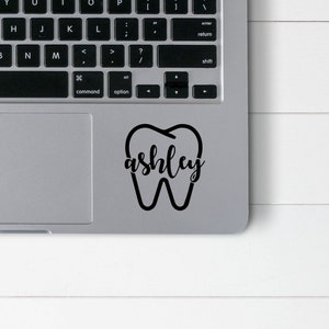 Puede incluir: Pegatina de vinilo en blanco y negro de un diente con el nombre "ashley" en su interior. La pegatina está en un portátil plateado.