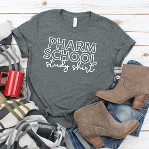 May include: A gray t-shirt with the text "PHARM SCHOOL study shirt" in white outlined letters. The shirt is laid out on a white wood surface with a blanket, a red mug, and brown ankle boots.