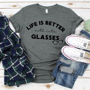 Puede incluir: Una camiseta gris con el texto "Life is better with cute glasses" y un gráfico de gafas. La camiseta está colocada sobre una superficie de madera con un par de zapatillas blancas y unos vaqueros azules.
