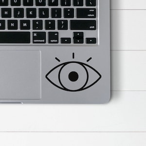 Puede incluir: Una pegatina de ojo en blanco y negro con rayos de luz que salen de ella. La pegatina está en una computadora portátil plateada.