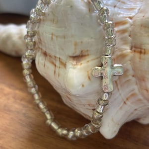 May include: A stretch bracelet composed of small, translucent clear glass beads and metallic silver-toned beads, featuring a single, central, polished silver-toned cross charm with a smooth, rounded surface and a simple, minimalist design.