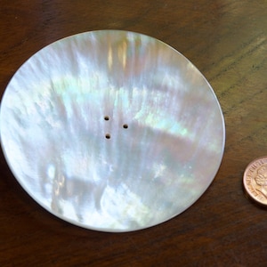 May include: A white iridescent mother of pearl dish with three small holes in the centre. The dish is round and has a slightly raised edge.