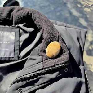 Puede incluir: Primer plano de una chaqueta negra con cuello de forro polar negro. Una piedra lisa, ovalada, de color marrón claro descansa sobre el cuello. La chaqueta tiene una cremallera y una etiqueta que dice "A MAN".