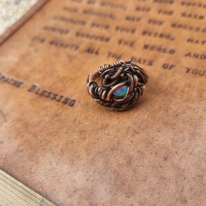 May include: A copper wire wrapped ring with an iridescent opal gemstone. The ring is sitting on a brown leather surface with text.