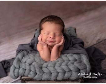 May include: A newborn baby is lying on a gray knitted blanket in a wooden box. The baby is smiling and has their hands under their chin. The baby is wearing a gray blanket.