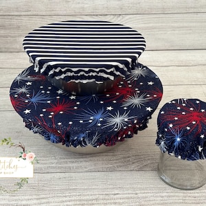 Op de afbeelding: Drie herbruikbare voedselhoezen in een blauw-wit gestreept patroon, een blauw-wit vuurwerkpatroon en een blauw-rood vuurwerkpatroon. De hoezen zijn gemaakt van stof en hebben een gerimpelde rand. De hoezen liggen op kommen en potten.