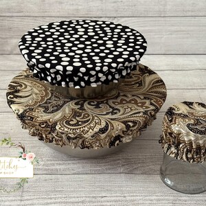 May include: Three reusable food covers in different sizes. The largest cover is black with white polka dots, the medium cover is brown with a paisley pattern, and the smallest cover is brown with a paisley pattern. The covers are on top of bowls and a jar.