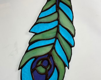 May include: A stained glass peacock feather with blue, green, and purple glass pieces. The feather is outlined in black and has a detailed eye design.