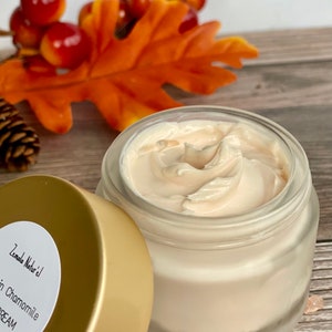 May include: A glass jar with a gold lid filled with a white cream. The jar has a label that reads "Zemaile Naturel" and "Chamomile Cream".