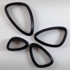 Può includere: Quattro oggetti neri di forma astratta sono disposti su uno sfondo bianco. Le forme hanno bordi arrotondati e un centro cavo, che assomigliano a forme di occhi stilizzate o forme geometriche. Gli oggetti variano leggermente in dimensioni.