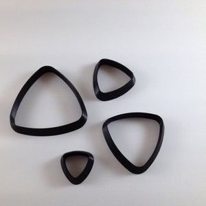 Può includere: Quattro formine per biscotti triangolari nere di diverse dimensioni sono disposte su uno sfondo bianco. Le formine hanno bordi arrotondati e una finitura liscia. Sembrano essere realizzate in plastica o materiale simile. Le forme sono semplici e moderne.