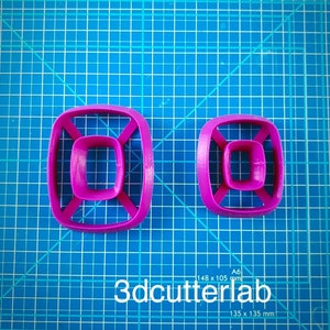 Puede incluir: Dos cortadores de galletas de color magenta con un diseño geométrico, cada uno con un cuadrado dentro de un cuadrado, conectados por barras. Los cortadores están sobre una esterilla de corte azul con líneas de cuadrícula. El texto "3dcutterlab" es visible.