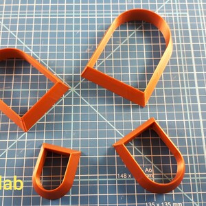 Puede incluir: Cuatro cortadores de galletas de color marr&oacute;n anaranjado en forma de U y de coraz&oacute;n, dispuestos sobre una esterilla de corte azul con cuadr&iacute;cula. Los cortadores son de diferentes tama&ntilde;os. El texto "3dcutterlab" es visible en la esquina inferior izquierda.