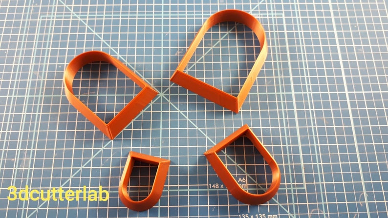 Puede incluir: Cuatro cortadores de galletas de color marr&oacute;n anaranjado en forma de U y de coraz&oacute;n, dispuestos sobre una esterilla de corte azul con cuadr&iacute;cula. Los cortadores son de diferentes tama&ntilde;os. El texto "3dcutterlab" es visible en la esquina inferior izquierda.