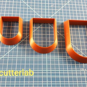 Puede incluir: Cuatro cortadores de galletas marrones en forma de U de diferentes tama&ntilde;os est&aacute;n dispuestos sobre una superficie de cuadr&iacute;cula azul. El cortador m&aacute;s peque&ntilde;o est&aacute; a la izquierda, el m&aacute;s grande a la derecha. El texto "3dcutterlab" es visible.