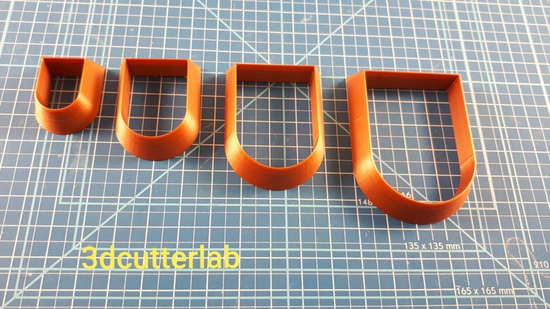 Puede incluir: Cuatro cortadores de galletas marrones en forma de U de diferentes tama&ntilde;os est&aacute;n dispuestos sobre una superficie de cuadr&iacute;cula azul. El cortador m&aacute;s peque&ntilde;o est&aacute; a la izquierda, el m&aacute;s grande a la derecha. El texto "3dcutterlab" es visible.